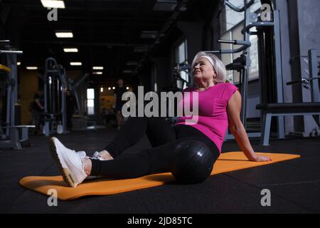 Entspannte ältere Frau, die auf einer Yogamatte sitzt und sich nach dem Training im Fitnessstudio ausruht Stockfoto