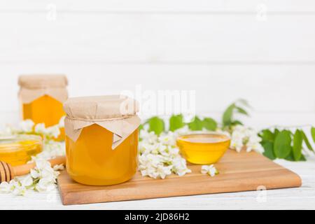 Süßes Honigglas umgab die Akazienblüten des Frühlings. Honig fließt aus einem Löffel in ein Glas. Gläser von klarem frischen Akazienhonig auf Holzgrund. Stockfoto