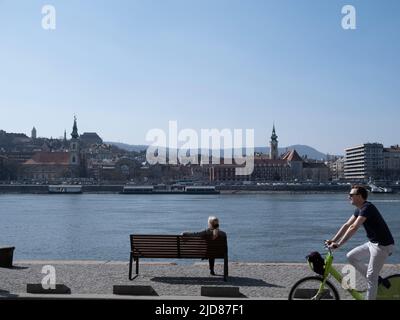 Budapest, Ungarn - März 2022: Einsame Frau sitzt auf der Bank und beobachtet die Donau und Mann radelt in Budapest, selektiver Fokus Stockfoto