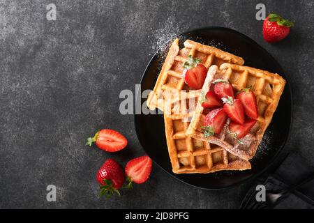 Belgische Waffeln. Hausgemachte Waffeln mit Erdbeeren, Puderzucker und eine Tasse Kaffee auf schwarzem Teller auf schwarzem Steintisch Hintergrund. Frühstück. Obere V Stockfoto