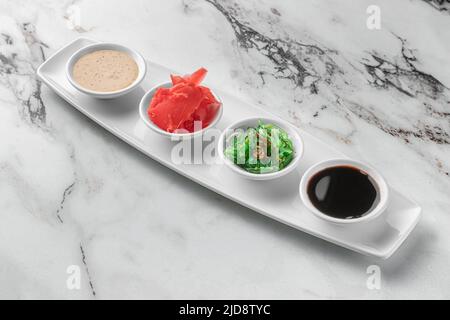 Set mit Soßen für Sushi auf weißem Teller, Ingwer, Soja, Sesam, Algen. Heller, strukturierter Marmorhintergrund, Seitenansicht Stockfoto