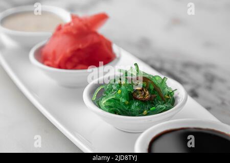 Set mit Soßen für Sushi auf weißem Teller, Ingwer, Soja, Sesam, Algen. Heller, strukturierter Marmorhintergrund, Seitenansicht, Nahaufnahme Stockfoto