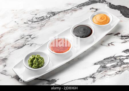 Set von Soßen für Sushi auf einem weißen Teller, Wasabi, würzig, kitschig, Soja. Heller strukturierter Marmorhintergrund, Seitenansicht Stockfoto