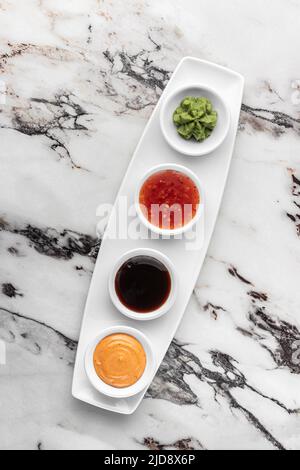 Set von Soßen für Sushi auf einem weißen Teller, Wasabi, würzig, kitschig, Soja. Heller strukturierter Marmorhintergrund, Draufsicht Stockfoto