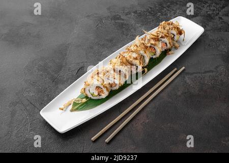 Set aus Bonito-Rollen mit gegrilltem Lachs, Avocado, Thunfisch-Sprünge und grünem Bambusblatt in einer weißen Keramikplatte mit Essstäbchen auf dunkelgrauer Struktur Stockfoto