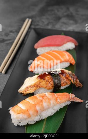 Set aus Nigiri mit Tiger Shrimp, tomago, Lachs, Thunfisch und grünem Bambusblatt in einer schwarzen Keramikplatte mit Essstäbchen auf einem dunkelgrauen strukturierten Hintergrund Stockfoto