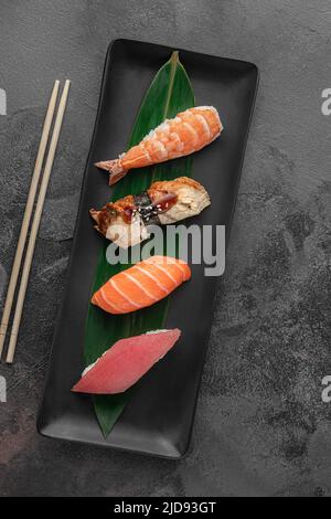 Set aus Nigiri mit Tiger Shrimp, tomago, Lachs, Thunfisch und grünem Bambusblatt in einer schwarzen Keramikplatte mit Essstäbchen auf einem dunkelgrauen strukturierten Hintergrund Stockfoto