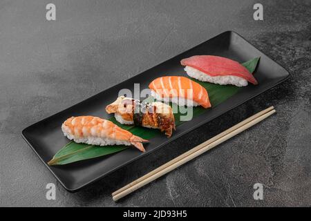 Set aus Nigiri mit Tiger Shrimp, tomago, Lachs, Thunfisch und grünem Bambusblatt in einer schwarzen Keramikplatte mit Essstäbchen auf einem dunkelgrauen strukturierten Hintergrund Stockfoto