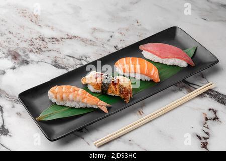 Set aus Nigiri mit Tiger Shrimp, tomago, Lachs, Thunfisch und grünem Bambusblatt in einer schwarzen Keramikplatte mit Essstäbchen auf einem hellweißen strukturierten Marmor Stockfoto