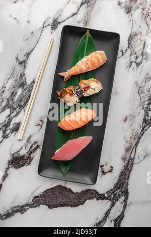 Set aus Nigiri mit Tiger Shrimp, tomago, Lachs, Thunfisch und grünem Bambusblatt in einer schwarzen Keramikplatte mit Essstäbchen auf einem hellweißen strukturierten Marmor Stockfoto