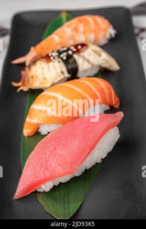 Set aus Nigiri mit Tiger Shrimp, tomago, Lachs, Thunfisch und grünem Bambusblatt in einer schwarzen Keramikplatte mit Essstäbchen auf einem hellweißen strukturierten Marmor Stockfoto