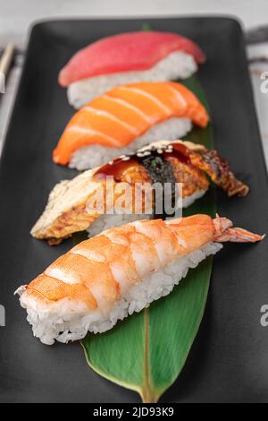 Set aus Nigiri mit Tiger Shrimp, tomago, Lachs, Thunfisch und grünem Bambusblatt in einer schwarzen Keramikplatte mit Essstäbchen auf einem hellweißen strukturierten Marmor Stockfoto