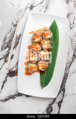 Set aus heißen gebackenen Brötchen mit Lachs, Teriyaki, Nori, Reis und grünem Bambusblatt in einer weißen Keramikplatte mit Essstäbchen auf einem hell strukturierten Marmorbac Stockfoto