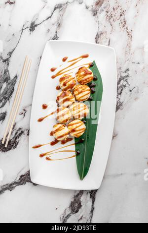 Set von heiß gebackenen ricco-Rollen mit Garnelen, Mozzarella, Nori, Reis und grünem Bambusblatt in einer weißen Keramikplatte mit Essstäbchen auf einem hellen texturierten ma Stockfoto