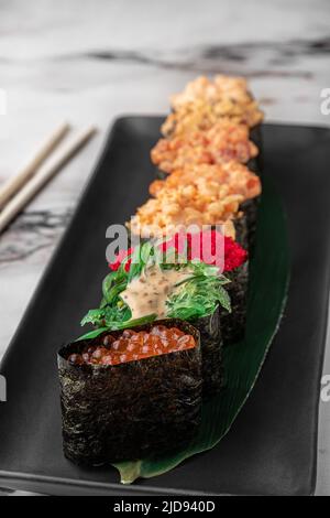 Set aus Gunkan mit Lachs, chuka-Salat, fliegendem Fischrogen, Thunfisch und grünem Bambusblatt in einer schwarzen Keramikplatte mit Essstäbchen auf einer hellweißen Struktur Stockfoto