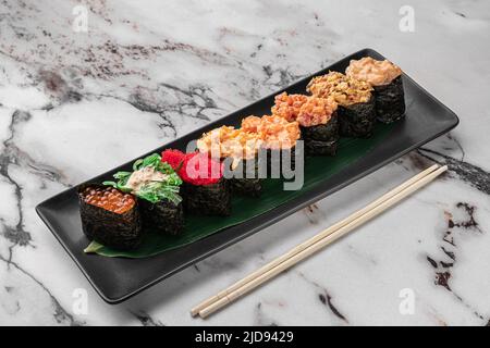 Set aus Gunkan mit Lachs, chuka-Salat, fliegendem Fischrogen, Thunfisch und grünem Bambusblatt in einer schwarzen Keramikplatte mit Essstäbchen auf einer hellweißen Struktur Stockfoto