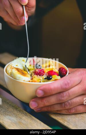 Nahaufnahme vertikale Foto männliche Hände halten eine Schüssel auf dem Holztisch, schöpfen Waffelschale voll mit Milch, Himbeere, Heidelbeeren, Mini-Waffeln und Stockfoto