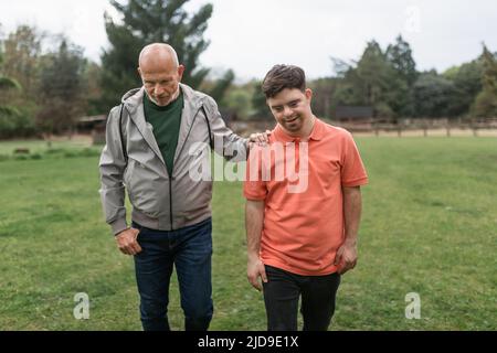 Glücklicher älterer Vater mit seinem jungen Sohn mit Down-Syndrom, der zusammen im Park läuft. Stockfoto