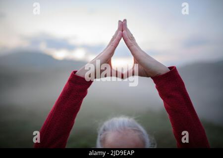 Ältere Frau macht am frühen Morgen Atemübungen in der Natur mit Nebel und Bergen im Hintergrund, Nahaufnahme Stockfoto