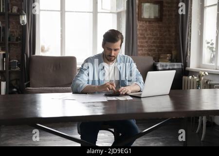 Ein junger, ernsthafter Mann, der am Schreibtisch sitzt, berechnet die Haushaltsfinanzen Stockfoto