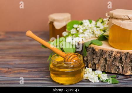 Süßes Honigglas umgab die Akazienblüten des Frühlings. Honig fließt aus einem Löffel in ein Glas. Gläser von klarem frischen Akazienhonig auf Holzgrund. Stockfoto