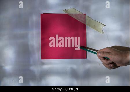 Die Hand eines reifen Mannes zeigt mit einem Bleistift auf ein Stück Papier. Leeres rotes quadratisches Stück Papier, das an der Wand befestigt ist. Eine abstrakte Licht- und Schattenzeichnung Stockfoto