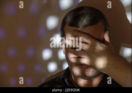 Ein Mann bedeckte sein Gesicht mit der Hand. Porträt eines Mannes mittleren Alters an einer Wand. Ein Licht- und Schattenmuster von sich wiederholenden Figuren an Wand und Th Stockfoto