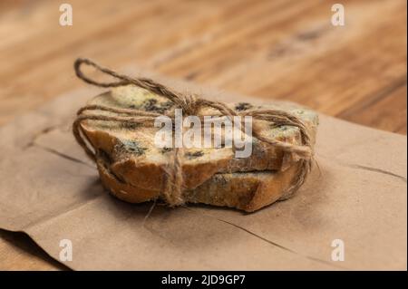 Schimmeliges Brot vor einem hölzernen Hintergrund. Zwei Scheiben Weizenbrot mit Pilzschimmel bedeckt. Die Scheiben sind mit Garn gebunden. Brauner Papierbeutel. Verdorben für Stockfoto