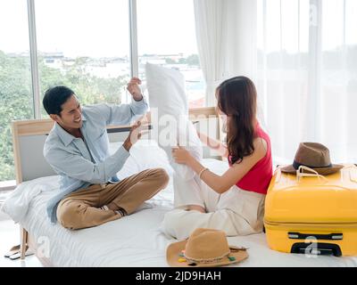 Paar Kissenschlacht. Asiatischer Mann im Denim-Hemd und schöne junge Frau, die zusammen mit Kissen neben dem gelben Koffer auf dem Bett sitzt und kämpft Stockfoto