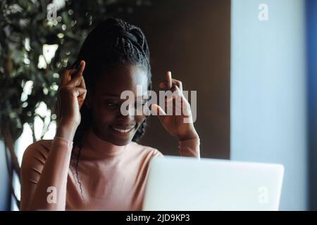 Lächelnde afroamerikanische Frau, die sich die Finger kreuzt und am Tisch im Kaffeehaus sitzt, von unten gesehen, verschwommener Vordergrund. Schwarzes Mädchen Freiberufler Stockfoto