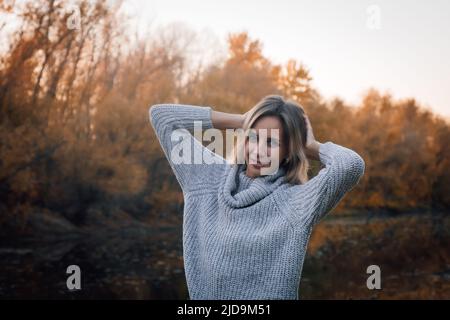 Nahaufnahme einer charmanten blonden Frau, die im Wald steht und lächelnd auf die Kamera blickt, wobei beide Hände hinter dem Kopf stehen und Bäume mit Sonnenuntergang bedeckt sind Stockfoto