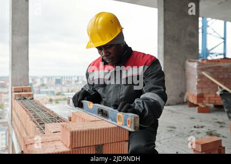 Reifer Baumeister in Arbeitskleidung und Schutzhelm halten Handwerkzeug auf Operation der Ziegelschicht Ebene, während die Ebenheit der Wand zu überprüfen Stockfoto