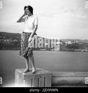1958, historisch, eine Frau in einem gepunkteten Rock, lächelnd, während sie für ein Foto auf einem Betonposten an einem Hafen an der Küste, England, Großbritannien, steht. Stockfoto