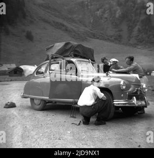 1958, historisch, Motortour durch die alpen, ein Mann, der auf der Bergstraße in Südtirol, Italien, einen Reifen wechselt. Gepäck unter einer wasserdichten Abdeckung auf dem Dach. Autopassagiere vor der Tür, die auf eine Karte schauen. Stockfoto