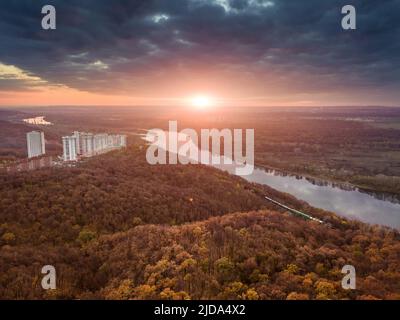 Landschaftlich reizvolle Aussicht auf die Eisenbahn in den Vororten der Stadt. Majestätische Sonnenuntergangswolken, die sich im Flusswasser spiegeln Stockfoto