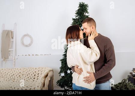 Schöne junge stilvolle Paar, Mann und Frau, in Strickpullover weiß und braun in den Armen des anderen auf dem Hintergrund des Weihnachtsbaums feiern Weihnachten zu Hause. Skandinavischer Minimalismus. Stockfoto