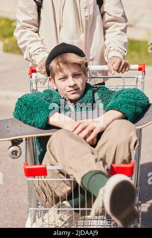 Teenager-Junge sitzt im Einkaufswagen mit seinem Skateboard und genießt das Reiten, während seine Freunde ihn Rollen Stockfoto