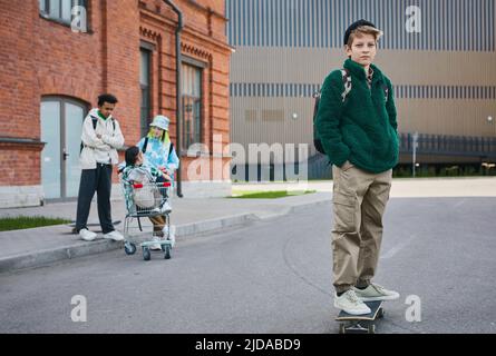 Porträt eines Jungen im Teenageralter, der auf dem Skateboard steht und die Kamera anschaut, während er mit seinen Freunden Zeit im Freien verbringt Stockfoto