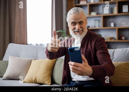 Lächelnder älterer Mann, der auf der Couch sitzt und den Daumen hochgibt, während er Computerspiele spielt, die Kamera anschaut und lächelt. Stockfoto