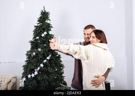 Fröhliches junges stilvolles Paar, tanzende Männer und Frauen, in Strickpullover weiß und braun in den Armen des anderen auf dem Hintergrund des Weihnachtsbaums, der zu Hause Weihnachten feiert. Skandinavischer Minimalismus. Stockfoto