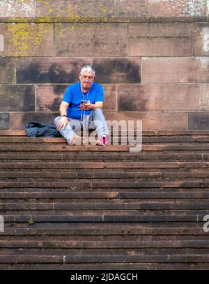 Ein ehrliches Foto eines Mannes, der mit seinem Smartphone auf den Stufen der Liverpool Cathedral sitzt und sich konzentriert, wobei seine Zunge etwas herausgestreckt ist. VEREINIGTES KÖNIGREICH Stockfoto