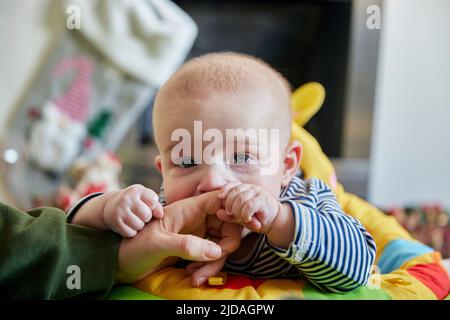 Nahaufnahme eines sechs Monate alten Jungen, der sich am Finger der Mutter hält und die Kamera anschaut Stockfoto