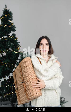 Junge schöne Mädchen hält Korbkorb auf dem Hintergrund der geschmückten Weihnachtsbaum mit Girlanden. Frau in weißem Strickpullover. Minimalistisches Konzept von Weihnachten und Neujahr. Kopierbereich, vertikal Stockfoto