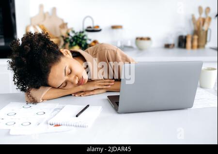 Chronischer Schlafentzug. Erschöpftes afroamerikanisches Mädchen, freiberufliche Mitarbeiterin oder Studentin, schlief auf dem Desktop in der Nähe des Laptops ein, überfordert von der Arbeit oder dem Online-Studium, braucht Ruhe und Schlaf Stockfoto