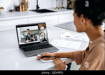 Über der Schulter Blick auf afroamerikanische Mädchen von Laptop-Bildschirm mit multirassischen Menschen. Brainstorming von Frauen mit Arbeitskollegen, Hören von Online-Webinaren mit Laptop und Videokonferenz Stockfoto