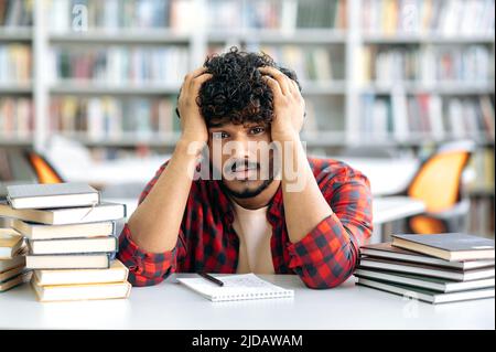 Müde erschöpfter inder oder araber, in lässiger Kleidung, sitzt ein Universitätsstudent oder ein Gymnasiast an einem Schreibtisch in einer Bibliothek zwischen Büchern, hält seine Hände auf dem Kopf, sieht vom Lesen müde aus Stockfoto