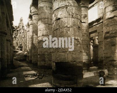 Dieses Bild aus dem Jahr 1910 zeigt die Ruinen der Säulenhalle in Karnak. Die große Hypostyelhalle Karnak besteht aus 134 riesigen Sandsteinsäulen in Form von Papyrus-Stielen. Zwölf große Säulen in seinem zentralen Kirchenschiff sind über 70 Meter hoch und werden von riesigen, offenen Papyrus-Blütenkapitellen gekrönt. Die Ost-West-Hauptachse der Hypostyle-Halle wird von einer Doppelreihe mit 12 riesigen Säulen dominiert. Dieses Bild stammt aus einer Photogravure in Ägypten der deutschen Archäologen Ebers und Junghaendel. Stockfoto