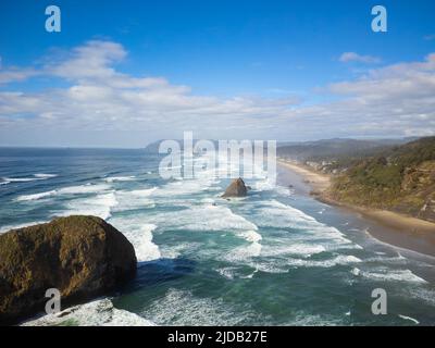 Wunderschöne Meereslandschaft. Türkisfarbenes Wasser mit hellweißen Wellen, ein großer Felsbrocken im Meer, eine sandige grüne Küste und ein strahlend blauer Himmel mit hellweißem c Stockfoto