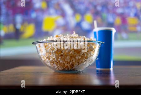 Ruhen Sie sich in der Nähe des Fernsehers aus, und schauen Sie sich Sportprogramme im Fernsehen an. Popcorn in einer großen Glasschüssel, trinken Sie in einem blauen Plastikglas. Der Hintergrund ist ein großer TV-Bildschirm wi Stockfoto