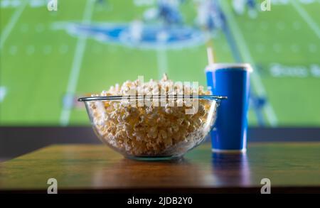 Fußballspiel auf Großbildfernseher. Auf dem Tisch steht eine Schüssel Popcorn und ein Getränk in einem Glas. Sehen Sie sich Ihre Lieblingssportprogramme im Fernsehen, Fußball und Amerika an Stockfoto